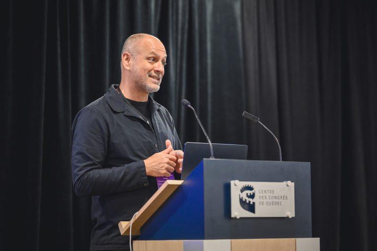 A presenter speaks from the podium during a session at the 1st International Congress on Safe Sport Research in Québec City, engaging participants with a smile.