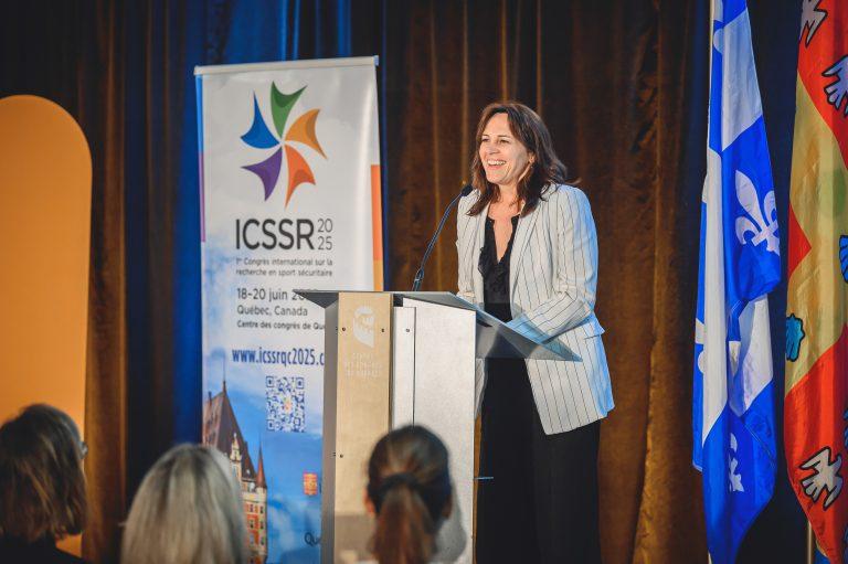 A speaker gives a keynote address at the 1st International Congress on Safe Sport Research in Québec City, standing at a podium with the ICSSR 2025 banner in the background.