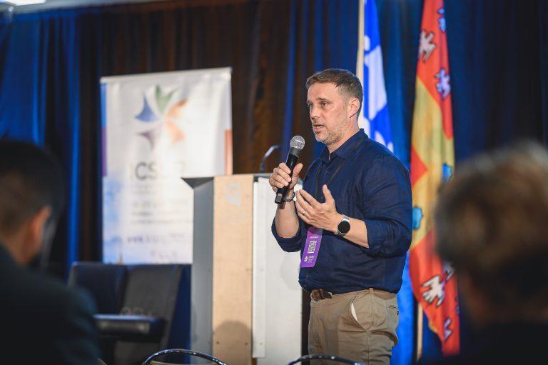 A presenter speaks into a microphone during a session of the 1st International Congress on Safe Sport Research, addressing participants gathered at the Québec City Convention Centre.