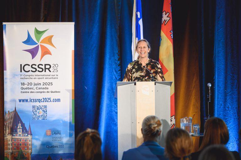 A speaker presents on stage during the 1st International Congress on Safe Sport Research in Québec City, standing at the podium before an attentive audience.