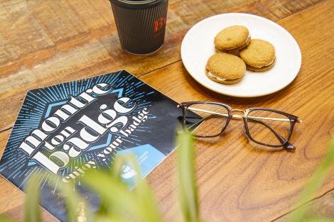 Photograph of the enjoy your badge card placed on a wooden table with some biscuits and a coffee cup